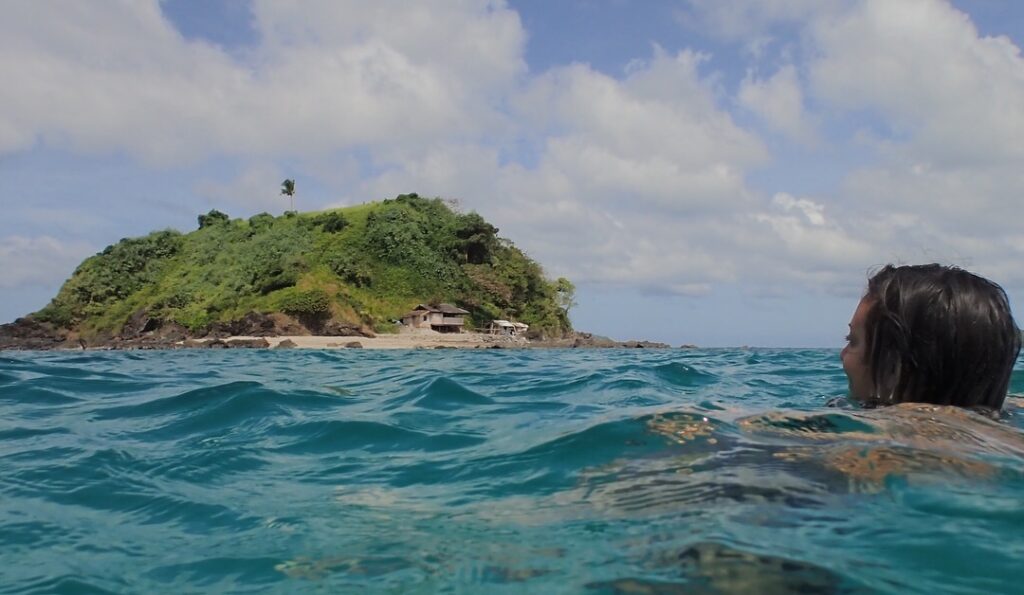 Brina, shown from behind, swimming in the blue waters of Nacpan Beach. Clouds and an island are in the background. Lesser known underrated travel destinations to add to your itinerary solo female travel