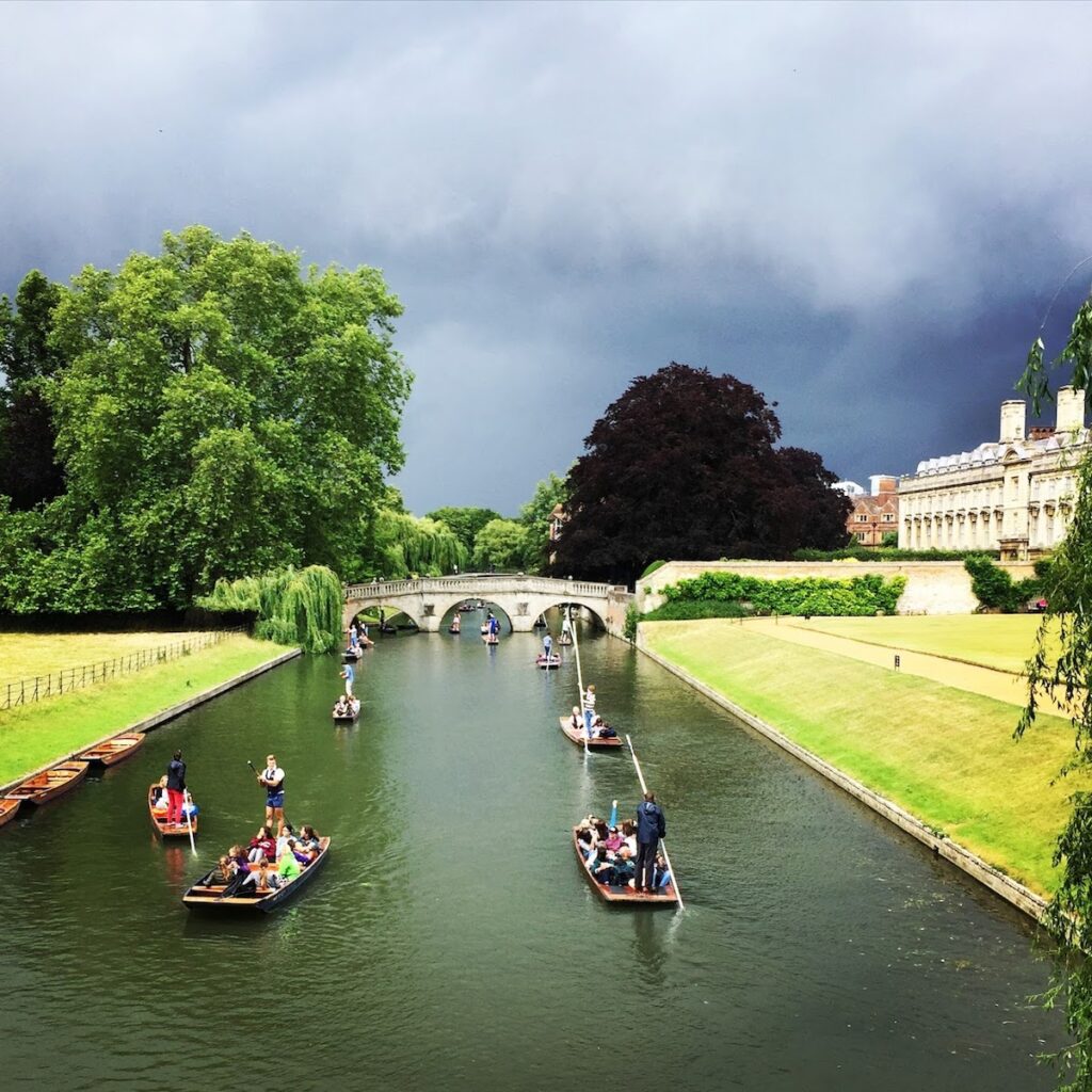 Punts in cambridge, with people sitting in them, cursing down the river with grey clouds in the distance. Lesser known underrated travel destinations to add to your itinerary solo female travel