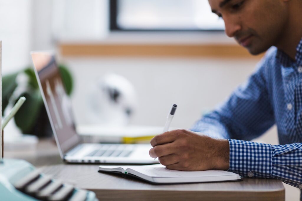 Man writing on piece of paper, with laptop open in front of him. resources freelance writer canva mailchimp newsletter